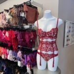 A mannequin in a red lace lingerie set is displayed next to racks of various colored lingerie pieces in a store.