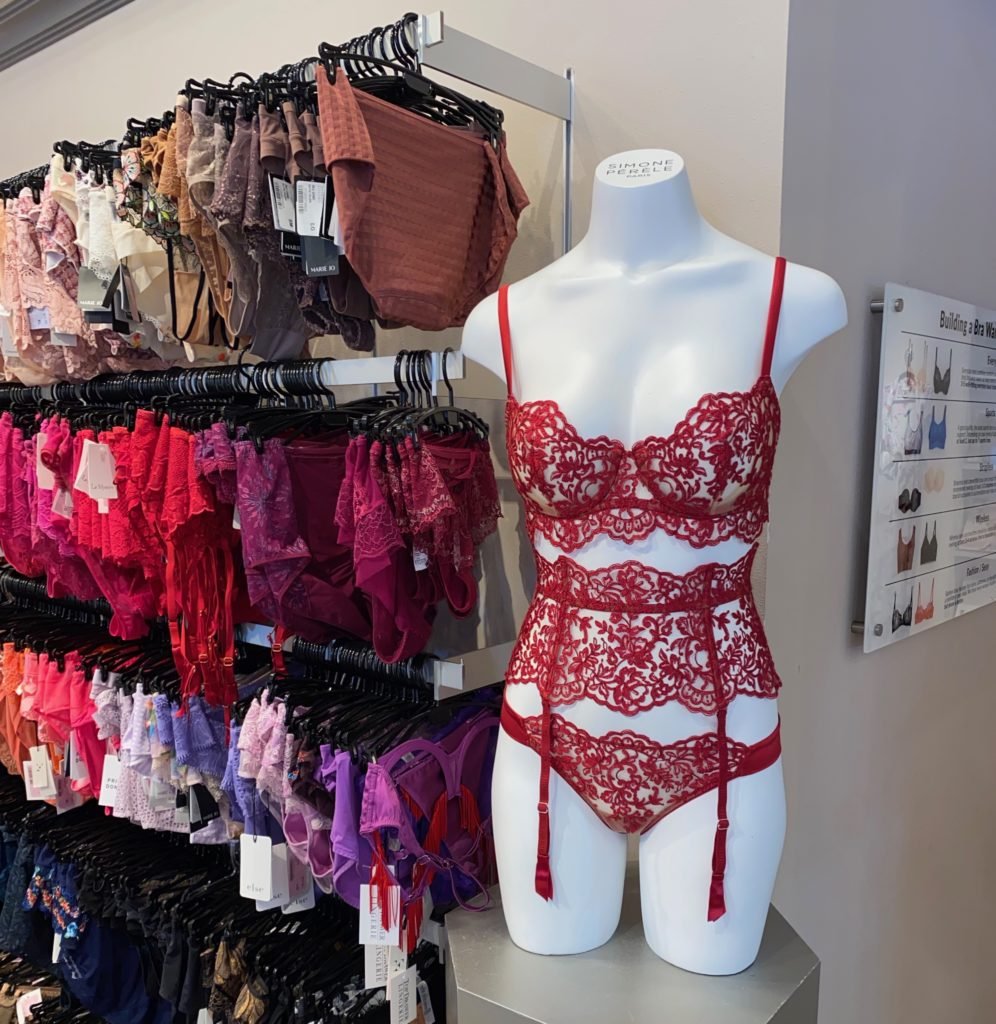 A mannequin in a red lace lingerie set is displayed next to racks of various colored lingerie pieces in a store.
